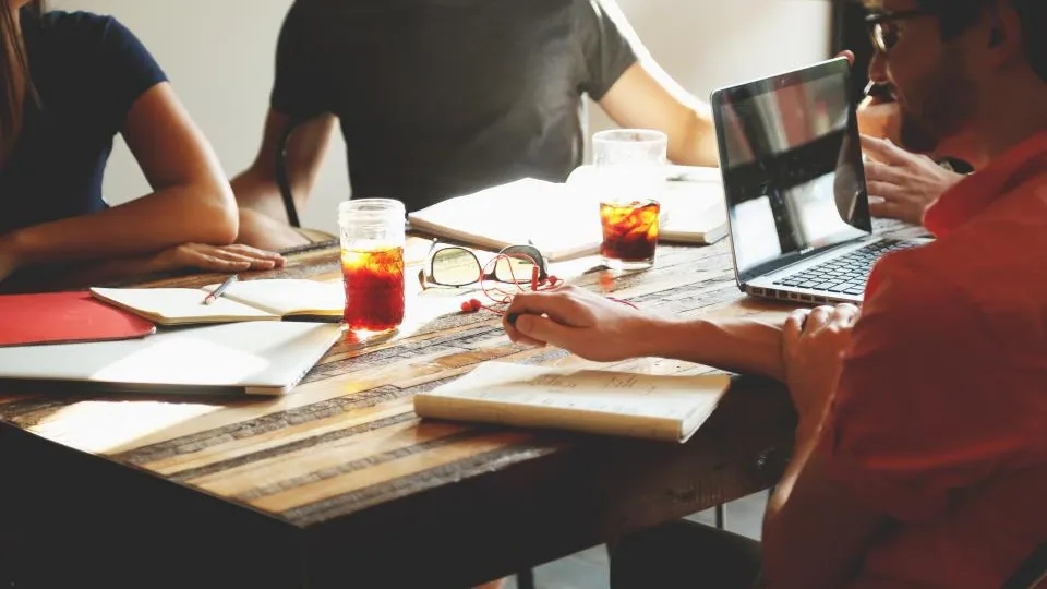 People talking around a wooden table with notebooks, drinks, and a laptop